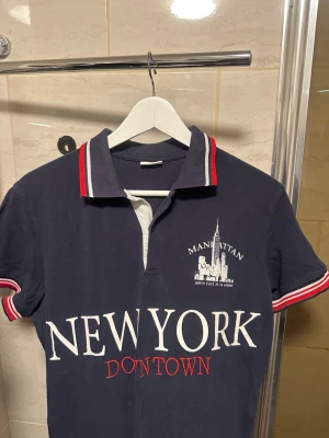 Marinblå pikétopp med New York-tryck - Marinblå pikétopp med vit och röd krage samt ärmslut. Stort tryck med texten 'NEW YORK DOWNTOWN' och Manhattan-motiv på bröstet. Klassisk pikékrage och kort ärm, perfekt för en sportig och urban look.