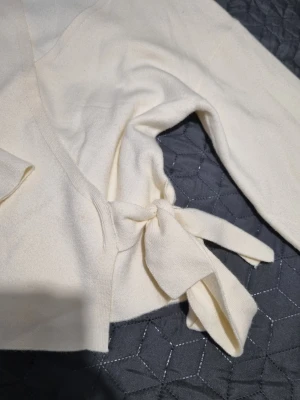 Krämvit omlottkofta med knytband - Snygg krämvit omlottkofta med långa ärmar och breda knytband i midjan. Koftan har en djup V-ringning och är tillverkad i ett mjukt, stickat material som känns skönt mot huden. Perfekt för lager på lager och enkel att styla till olika outfits. Bara provat ny utan prislapp I storlek L från Ginatricot tonåringsavdelning. 