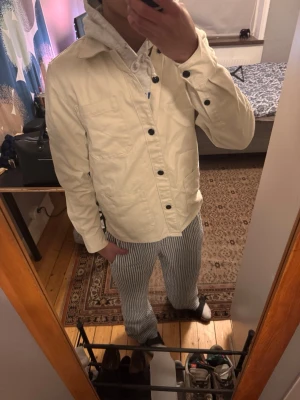 Beige overshirt med svarta knappar - Stilren beige overshirt med svarta knappar och tre fickor framtill. Klassisk krage och raka ärmar med knappdetaljer vid ärmslut. Perfekt lager-på-lager-plagg i bomull som funkar året runt.