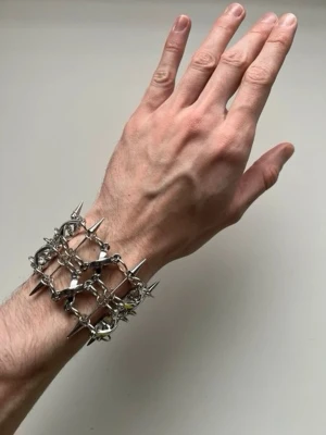 Silvrigt armband med nitar och kedjor - Unikt armband i silverfärgad metall med flera rader av kedjor och spetsiga nitar. Armbandet har ett rutnätsmönster och ger en edgy, alternativ look. Perfekt för dig som gillar statement-smycken och vill sticka ut med din stil.