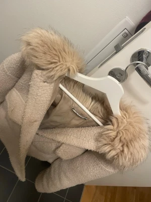 Chelsea kappa - Supermjuk och trendig beige teddyjacka med fluffig fuskpäls på kragen.