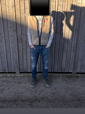 Beige fleeceväst från Patagonia - Säljer en beige fleeceväst från Patagonia med marinblå dragkedja och röda detaljer. Västen har två fickor med dragkedja och klassisk logga på bröstet. Perfekt för lager-på-lager och chill stil. Använd i cirka 1 månad.