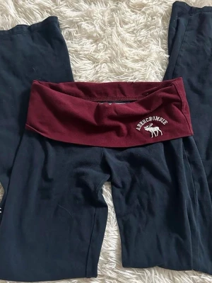 Svarta leggings med vinröd midja - Snygga svarta leggings från Abercrombie med bred vinröd midjeresår och vit logga med älg. Perfekta för en avslappnad och trendig look. Mjukt och stretchigt material som sitter skönt hela dagen.