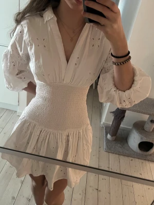Vit kort klänning med broderade detaljer - Superfin vit kortklänning med broderade hål och volangkant. Klänningen har v-ringning, markerad midja med smock och puffiga långärmar. Perfekt för sommarens alla tillfällen och ger en romantisk vibe. Köpte på plick men aldrig använt och den förra ägaren hade bara använt den en gång!!! 