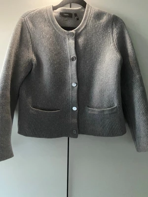 Grå kofta från Theory med knappar - Snygg grå kofta från Theory med rund halsringning och silverfärgade knappar framtill. Koftan är stickad i ett mjukt material och har två fickor framtill. Perfekt att slänga på sig över en t-shirt eller skjorta.