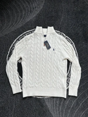 Vit kabelstickad tröja Polo Ralph Lauren - Vit kabelstickad tröja från Polo Ralph Lauren med halv dragkedja och hög krage. Tröjan har det klassiska broderade logotypen på bröstet och ribbade muddar vid ärmslut och nederkant. Perfekt för en stilren look.