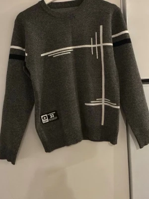 Mörkgrå stickad tröja med grafiskt mönster - Snygg mörkgrå stickad tröja med vita och svarta grafiska linjer på framsidan. Tröjan har rund hals, långa ärmar och en patch med text nertill. Perfekt för dig som gillar en modern och stilren look.