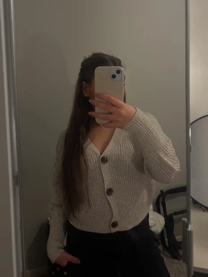 Beige stickad kofta - Säljer en beige stickad kofta från Vero Moda med v-ringning och stora bruna knappar framtill. Koftan är kortare i modellen, perfekt att slänga över en t-shirt för en chill look.