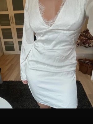 Vit kort klänning med spetsdetaljer - Säljer en vit kort klänning med v-ringning och spetsdetaljer från nakd. Klänningen har lång ärm och är gjord i ett glansigt material som ger en elegant look. Perfekt för student eller liknande💕 strl 36