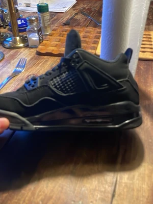Svarta Air Jordan 4 sneakers - Snygga helsvarta Air Jordan 4 sneakers med meshpaneler på sidorna, synlig Air-dämpning i sulan och klassisk Jumpman-tag. Skorna har en robust siluett, högt skaft och detaljer i mocka och läder. Perfekta för dig som gillar streetwear och ikoniska modeller.skorna är i storlek 39 