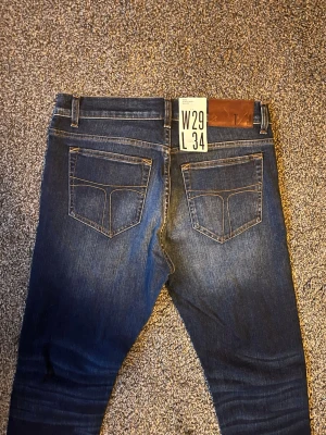 Mörkblå jeans från Tiger of Sweden - Snygga mörkblå jeans från Tiger of Sweden med klassisk femficksmodell och kontrastsömmar. Jeansen har en rak passform och är tillverkade i ett slitstarkt denimtyg. Perfekta för dig som gillar stilrena och tidlösa jeans.