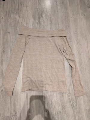 Beige ribbad offshoulder topp - Trendig beige offshoulder topp med långa ärmar och ribbad struktur. Perfekt att styla med jeans eller kjol för en clean och snygg look. Mjuk och stretchig känsla i materialet.