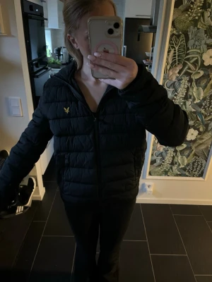 Svart pufferjacka från Lyle & Scott - Säljer en svart pufferjacka från Lyle & Scott med gul logotyp på bröstet. Jackan har dragkedja framtill, hög krage och quiltad design. Perfekt för kalla dagar och har en modern, sportig look. Säljer på grund av att jackan är för stor 