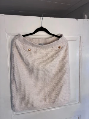 Stickad vit/beige kjol från Holly & Whyte - Säljer en stickad vit/beige kjol från Holly & Whyte med snygg struktur och två dekorativa guldfärgade knappar framtill. Kjolen har en rak, kort modell och bred resår i midjan för skön passform. Perfekt att matcha med en enkel topp eller stickad tröja. Passar S/M