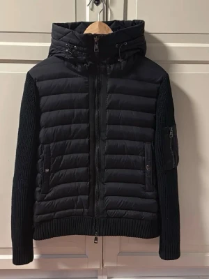 Svart moncler pufferjacka med stickade ärmar - Snygg svart pufferjacka med quiltad front och huva, samt stickade ärmar och rygg. Jackan har dragkedja framtill, två fickor med knappar och en extra ficka med dragkedja på ärmen. Perfekt för kalla vinterdagar och riktigt trendig look.