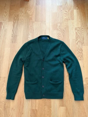 Polo Ralph Lauren Cardigan - Tja! Säljer nu en stilren mörkgrön cardigan från Polo Ralph Lauren i otroligt fint skick,  fem knappar framtill och i frägen mörkgrön. Nypris ca 3000 kr, säljer för knappt halva! Två fickor framtill och ribbade muddar vid ärmslut och nederkant. Perfekt för lager på lager och enkel att matcha. Storlek M. Pris går alltid att diskuteras✅. 