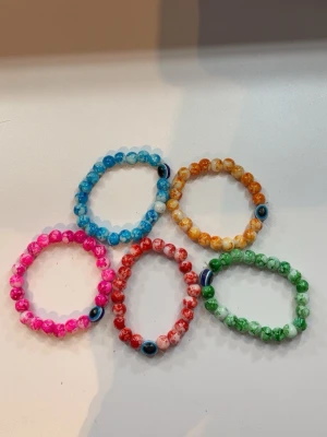 Färgglada pärlarmband med öga - Fem elastiska armband med färgglada pärlor i blått, orange, rosa, rött och grönt. Varje armband har ett pärlformat öga som detalj. Perfekta för att ge din stil en lekfull och trendig touch.