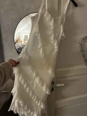 Vit volangklänning med smala axelband - Supersöt vit klänning med smala axelband och flera diagonala volangdetaljer. Klänningen har en lätt figurnära passform och är perfekt för sommarens alla tillfällen. Materialet känns mjukt och svalt, troligen polyester eller bomull.