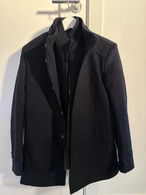 Mörkblå ullrock med innerväst - Stilren mörkblå rock med klassisk krage och dold dragkedja samt knappar. Rocken har två sidofickor, innerfickor och innerväst. Bara provat. Liten i storlek.