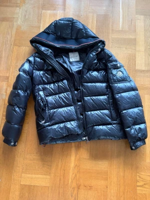 Svart dunjacka från Moncler med huva - Snygg svart dunjacka från Moncler med glansig finish och stor huva. Jackan har quiltad design, dragkedja framtill, två sidofickor och Moncler-logga på ärmen. Perfekt för kalla vinterdagar och riktigt trendig look. Storlek 2 vilket motsvarar M. Pris går att diskutera!🤩