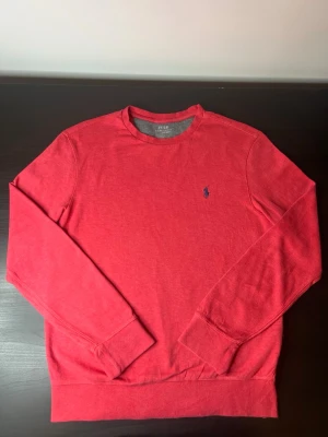 Röd sweatshirt från Polo Ralph Lauren - ÄKTA Polo Ralph Lauren sweatshirt i snygg röd färg. Klassisk och tidlös design med broderad logga på bröstet. Mjuk och bekväm kvalitet med rund hals och ribbade muddar – perfekt för både vardag och en mer uppklädd casual look.