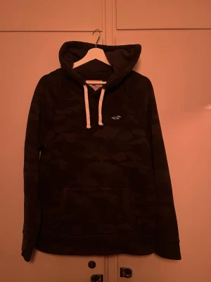 Camo hoodie från Hollister - Mörkgrå hoodie från Hollister med camomönster och vita dragsnören i huvan. Klassisk känguruficka framtill och broderad logga på bröstet. Perfekt för en avslappnad streetwear-look.