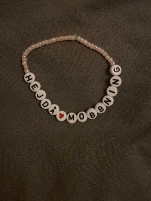 Pärlarmband med text Hejda Mobbning - Sött pärlarmband med vita och rosa pärlor, där de vita pärlorna har svarta bokstäver som bildar texten 'HEJDÅ MOBBNING' och ett rött hjärta. Armbandet är elastiskt och har en ungdomlig vibe.