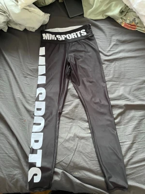 Svarta leggings från MM Sports - Svarta leggings från MM Sports med vit logga längs ena benet och bred resår i midjan med tryckt text. Tight passform och stretchigt material som passar perfekt till träning eller chill. Snygg och sportig look med hög midja. Använda väldigt fåtal gånger.