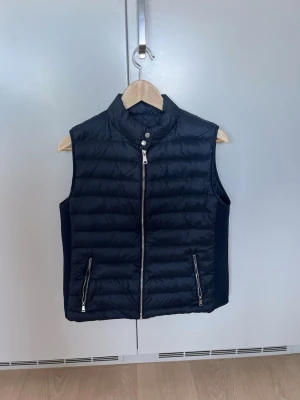 Svart dunväst med dragkedja - Snygg svart dunväst med ståkrage och två dragkedjefickor framtill. Västen har quiltad design och stängs med både dragkedja och tryckknapp vid halsen. Perfekt för lager-på-lager och enkel att matcha med olika outfits. Storlek Xl på papper men sitter som S