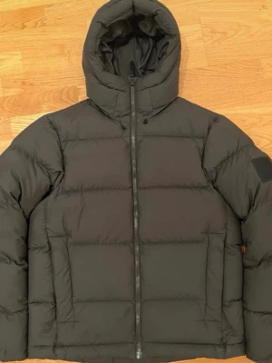 Grön pufferjacka med huva - Säljer en svart pufferjacka med huva och dragkedja framtill. Jackan har en stilren look med breda, horisontella kanaler och en patch på ena ärmen. Perfekt för kalla dagar och riktigt skön att bära. Passar dig som gillar minimalistisk och modern stil.