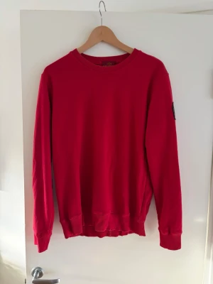 Röd sweatshirt från Parajumpers - Röd sweatshirt från Parajumpers med rund halsringning och ribbade muddar vid ärmslut och nederkant. Klassisk patch med PJS-logga på vänster ärm. Perfekt för en avslappnad och stilren look. Storlek M. Pris går att diskutera!🤩