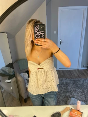 Beige axelbandslös broderad tubtopp - Söt beige axelbandslös tubtopp med broderade hålmönster och volangkant. Toppen har en bandeauformad överdel och är perfekt till jeans eller kjol för en avslappnad och trendig look.