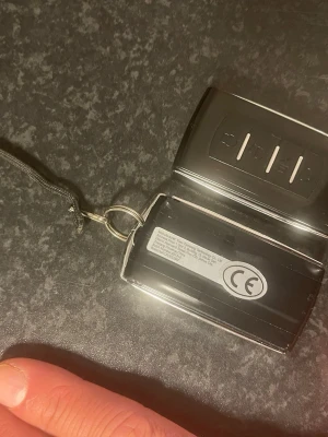 Svart bilnyckel med nyckelring - Elektronisk svart bilnyckel med tre knappar och CE-märkning. Nyckeln har en metallring och ett svart band för enkel upphängning. Tillverkad i plast och metall, stilren och praktisk design.