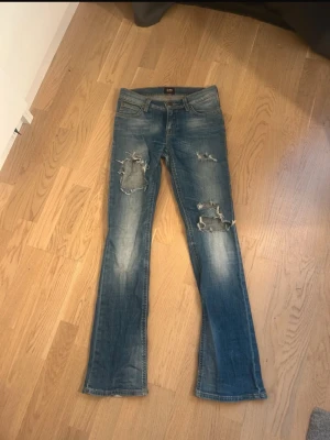 Blå slitna jeans från Lee - Säljer ett par blå jeans från Lee med slitningar och hål på båda benen. Jeansen har klassisk femficksmodell, låg midja och lätt utsvängda ben. Materialet är denim i bomull och färgen är tvättad blå med ljusare partier. Perfekta för en avslappnad och trendig look.