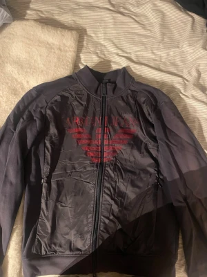  Zip-hoodie från Armani Jeans - Snygg svart hoodie från Armani Jeans med dragkedja och stort rött logotryck på bröstet. Tröjan har hög krage och ribbade muddar. Nypris ligger på typ 2000kr men jag säljer den för 600kr. 