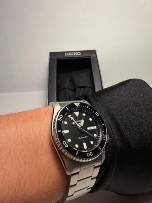 Seiko automatisk klocka i svart/silver - Full set med kvitto - Stilren Seiko automatisk klocka med svart urtavla och tydliga vita index. Klockan har dag- och datumvisning samt en robust länk i silverfärgat stål. Perfekt för dig som gillar klassisk och sportig design. Extra länkar, orginalbox och kvitto medföljer.