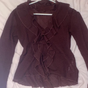 Vinröd volangblus från Gina Tricot - Säljer en vinröd blus från Gina Tricot med långa ärmar och volangdetaljer längs hela framsidan och kragen. Blus en är tunnstickad och har en feminin, figurnära passform. Perfekt för dig som gillar romantiska detaljer och mjuka material.