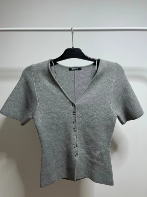 Grå kortärmad kofta från Gina Tricot - Stilren grå kofta från Gina Tricot med korta ärmar och v-ringning. Koftan har en figurnära passform och knäpps med små silverfärgade knappar, andvänd 1 gång