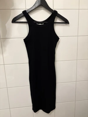 Svart ribbad klänning från H&M - En svart, ärmlös ribbad klänning från H&M i figurnära modell. Klänningen är kort och har en enkel, stilren design med rund halsringning. Perfekt för dig som gillar minimalistisk stil och vill ha något bekvämt och snyggt.