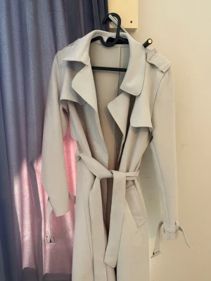 Ljusbeige trenchcoat med knytskärp - Stilren ljusbeige trenchcoat med klassisk krage, knytskärp i midjan och dekorativa band vid ärmsluten. Jackan har en lång, rak passform och snygga detaljer framtill. Perfekt för dig som vill ha en tidlös och trendig look.