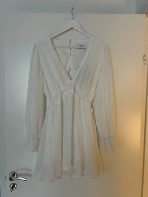 Vit kortklänning med volang och öppen rygg - Supersöt vit kortklänning från NA-KD med v-ringning, volangdetaljer och långärmad design. Klänningen har ett diskret rutmönster, markerad midja och öppen rygg med knappstängning i nacken. Perfekt för studenten 