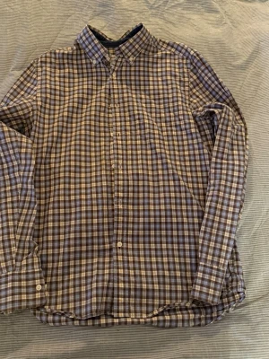 Rutig skjorta från Peak Performance - Rutig skjorta från Peak Performance i brunt, blått och vitt med klassisk krage och knäppning framtill. Skjortan har lång ärm och bröstficka, perfekt för en avslappnad men snygg stil.