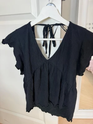 Svart v-ringad topp med knyt - Svart topp med v-ringning och knyt i nacken. Modellen har korta ärmar och en lätt volang nedtill. 