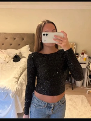 Svart glittrig croppad topp - Säljer en svart långärmad topp med glittriga paljetter och öppen rygg. Modellen är croppad och har en tight passform som framhäver midjan. Perfekt för dig som vill sticka ut med lite extra glitter.