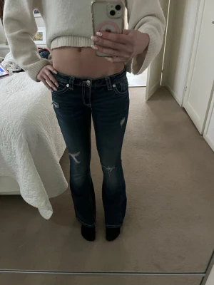 Mörkblå bootcut jeans med slitningar - Säljer ett par mörkblå bootcut jeans med slitna detaljer och kontrastsömmar. Jeansen har låg midja, fem fickor och snygg broderad bakficka. Perfekta för en avslappnad och trendig look.