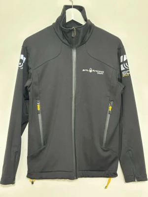 Svart vindjacka från Sail Racing - Svart vindjacka från Sail Racing med hel dragkedja, höga krage och tryckta loggor på bröstet och ärmarna. Jackan har två fickor med dragkedja och gula detaljer samt justerbar nederkant. Perfekt för en sportig och stilren look.