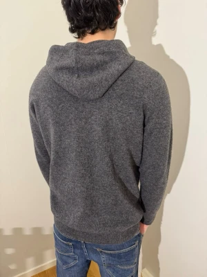 Mörkgrå hoodie i mjuk ullmix - Mysig mörkgrå hoodie med dragsko och huva, tillverkad i en mjuk och varm ullmix. Klassisk passform med ribbade muddar vid ärmslut och nederkant. Perfekt för en chill och stilren look. 