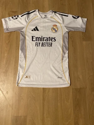  Vini Jr fotbollströja  - Snygg vit Real Madrid matchtröja från Adidas med Vini Jr. och nummer 7 på ryggen. Tröjan har svarta detaljer, klubbmärke på bröstet, Champions League-märke på ärmen och sponsortryck framtill. Tillverkad i lätt och ventilerande material, perfekt för fotboll.