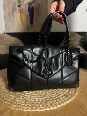 Svart quiltad YSL handväska i skinn - Snygg svart handväska från YSL i quiltat skinn med kedjehandtag och präglad logga framtill. Väskan har en mjuk, strukturerad form och stilrena detaljer i svart metall. Perfekt för dig som vill ha en lyxig och trendig accessoar.