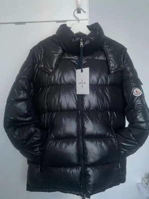 Svart dunjacka från Moncler - Säljer en svart, glansig dunjacka från Moncler med hög krage och dragkedja framtill. Jackan har två fickor med dragkedja och klassisk Moncler-logga på ärmen (Loggan funkar att skannas). Perfekt för kalla vinterdagar och riktigt snygg streetstil. Priset kan diskuteras!!